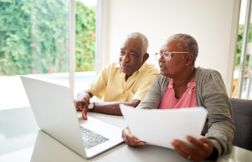 A senior living couple is sitting down at a table together, researching senior living options on their laptop